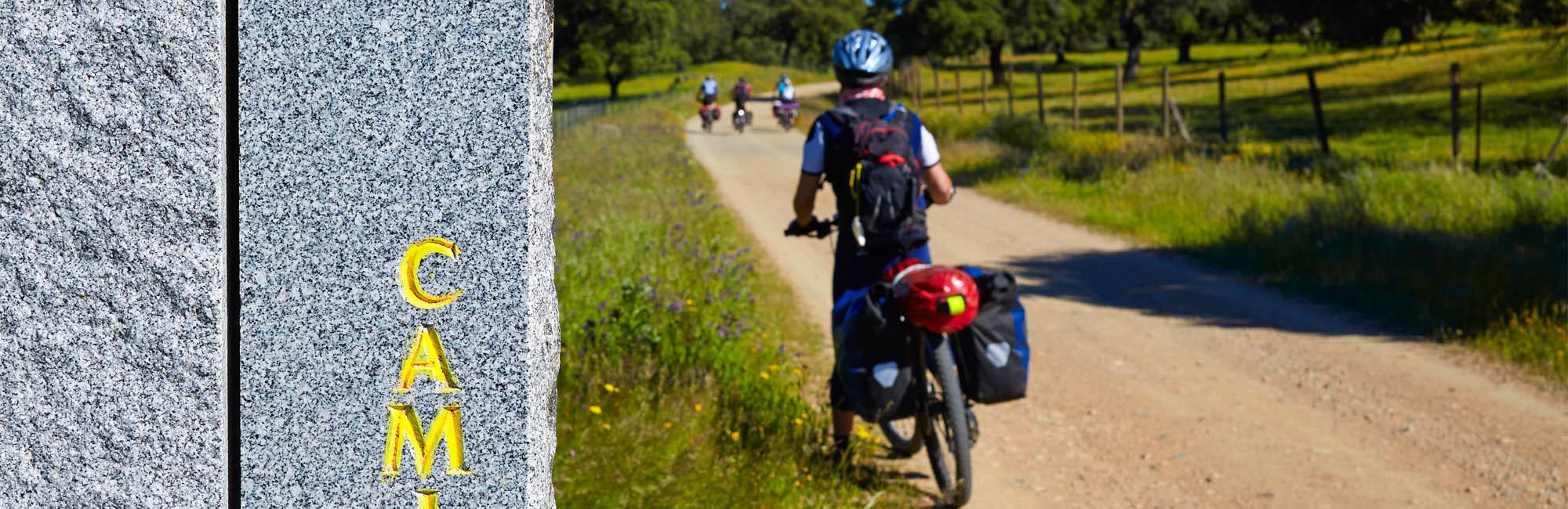 Sulla sinistra una pietra con inscrizione gialla, sulla destra un uomo fermo in bicicletta, carico di 2 borse laterali blu che guarda avanti verso i suoi compagni di cammino